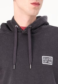 Felpa con cappuccio in tessuto grigio scuro con finitura testurizzata, dotata di un patch con logo frontale e lacci con punte in metallo.