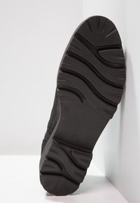 Suela de zapato negra con surcos profundos y ondulados, colocada en vertical contra una pared blanca de esquina, tamaño marcado como 37 en la suela.