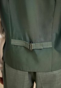 Grüne taillierte Jacke mit texturiertem Stoff, einem metallischen Schnallendetail und passenden Hosen mit dezentem Karomuster.