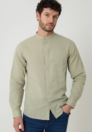 Hombre con cabello rizado y oscuro, vistiendo una camisa de botones con cuello mandarín de color verde claro y jeans azules, con la mano en el bolsillo, sobre un fondo neutro.