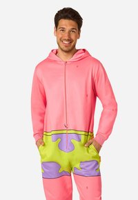 Lachende man die een kleurrijk zip-up hoodie en broekset draagt met roze, groene en paarse abstracte patronen, handen in zakken.