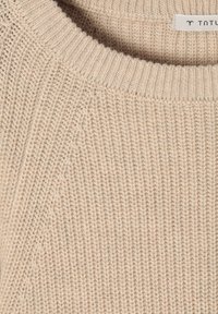 Beżowy sweter z prążkowanego materiału z okrągłym dekoltem, z ukośnymi i pionowymi wzorami ściegów oraz małą białą metką z logo marki wewnątrz kołnierza.