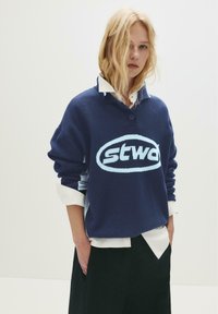 Blonde Frau mit lockigem Haar trägt einen navyblauen Pullover mit weißem Logo über einem weißen Hemd mit Kragen und steht mit den Händen in den Taschen ihrer schwarzen Hose.