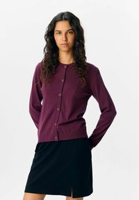 Cardigan violet avec un col rond, des manches longues et une fermeture à boutons sur le devant, associé à une jupe noire avec une fente sur le côté.