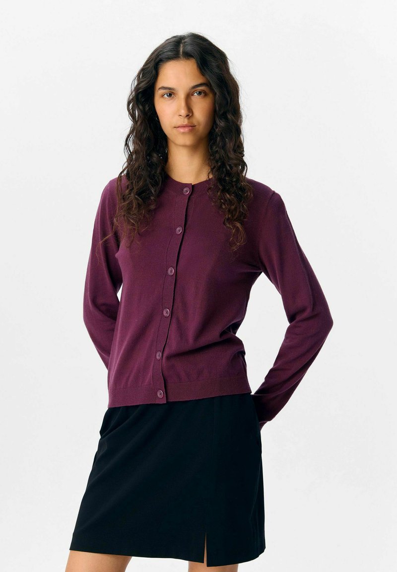 Cardigan violet avec un col rond, des manches longues et une fermeture à boutons sur le devant, associé à une jupe noire avec une fente sur le côté.