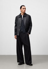Giacca di pelle nera con bottoni a pressione metallici sopra una camicia blu navy, abbinata a pantaloni neri larghi e scarpe nere. Texture liscia, design minimalista.