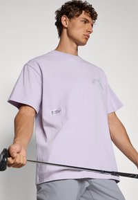 Ung mann i lavendel t-skjorte som holder en golfkølle horisontalt, ser til siden, mot en lys ensfarget bakgrunn.
