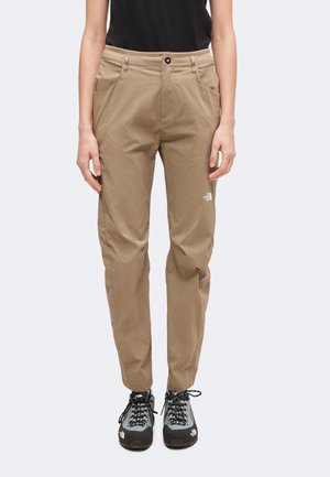 Persoon die beige North Face-broek, zwart T-shirt en grijs-zwarte wandelschoenen draagt, staat met ontspannen armen langs het lichaam.