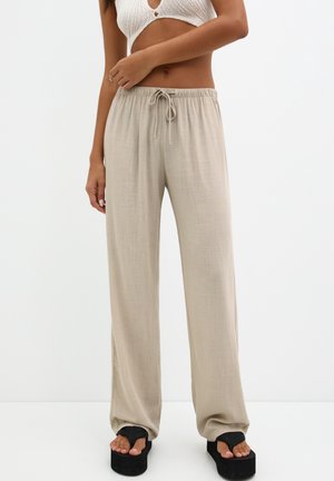 Frau trägt beige, weit geschnittene, mit Tunnelzug versehene Hosen, weißes, gestricktes Crop-Top und schwarze Plateau-Zehensandalen, steht vor weißem Hintergrund.