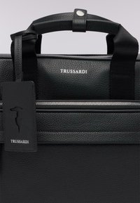 Svart läderportfölj med strukturerad yta, silverfärgad dragkedja och svarta tyghandtag. Har en tagg med logotyp som säger "TRUSSARDI."