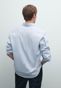 Jonge man met kort bruin haar, gekleed in een lichtblauw shirt met lange mouwen en opgerolde mouwen, en donkere broek, gezien van achteren.