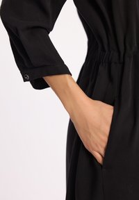 Czarna odzież o luźnym kroju, z marszczoną talią i podwiniętymi mankietami. Widoczne boczne kieszenie eksponują teksturę materiału.