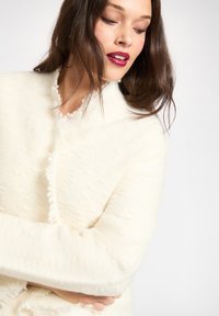 Witte textuurrijke jas van een zacht, pluizig materiaal. Heeft een kraagloos ontwerp en franjes aan de randen, om de armen van het model gewikkeld.