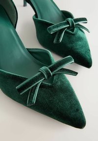 Et par mørkegrønne flade sko i velour med spidse tåer og dekorative velourbuer på en lys baggrund.