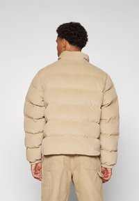 Beige korduroja puffer jaka ar vertikālām kanālu šuvēm, augstu apkakli un elastīgām aprocēm. Valkāta virs khaki biksēm, parādot teksturēto virsmu.