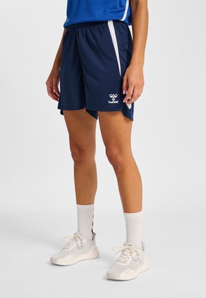 Person iført mørkeblå hummel sportsshorts, hvide sokker til midtpå læggen og hvide atletiksneakers mod ensfarvet baggrund.