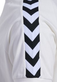 Hvid T-shirt med et sort og hvidt chevronmønsteret strikbånd langs skulderen. Blødt bomuldsstof med en afslappet pasform.