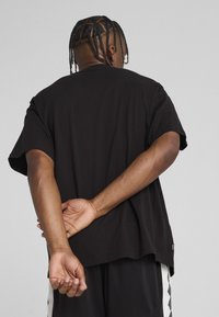 Sort bomulds t-shirt med korte ærmer, afslappet pasform, med buet kant og sideskilt; model står med hænderne foldet bag ryggen.