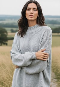 Jasnoszary, oversize sweter z prążkowanym, wysokim dekoltem, teksturowanym wzorem na dzianinie i bufkowymi rękawami, noszony z założonymi na siebie rękami.
