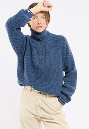 Femme portant un pull en maille bleu boutonné et un pantalon beige lève son bras, couvrant partiellement son front, avec une expression neutre.