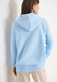 Femme portant un sweat à capuche bleu clair et un pantalon beige, debout de dos face à la caméra sur un fond clair uni.