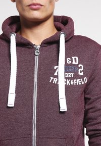Sudadera de tela burdeos oscuro con cremallera frontal y cordones blancos. Presenta el texto "TRACK & FIELD" y una superficie texturizada.