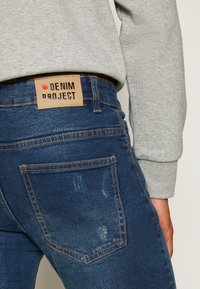 Tmavě modré džíny z denimu s opotřebovanou texturou a hnědou koženou etiketou s nápisem „DENIM PROJECT.“ Standardní zadní kapsy a viditelné oranžové švy.