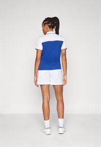 Camisola polo azul e branca, de mangas curtas e com colarinho, emparelhada com calções brancos. Tecido suave, corte atlético, poucos detalhes.