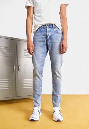 Personne portant un jean bleu clair, des baskets blanches avec des détails bleus et un t-shirt blanc, debout sur un sol jaune près de casiers métalliques gris.
