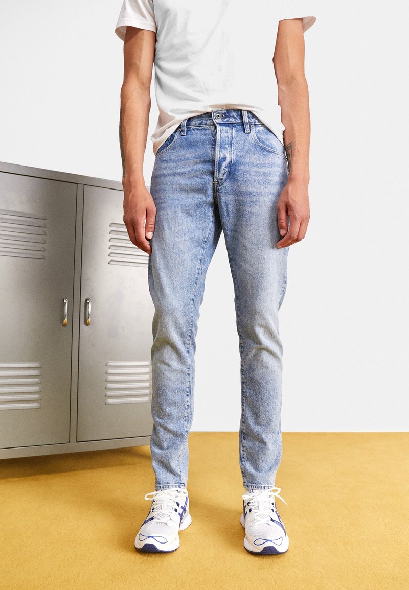 Personne portant un jean bleu clair, des baskets blanches avec des détails bleus et un t-shirt blanc, debout sur un sol jaune près de casiers métalliques gris.