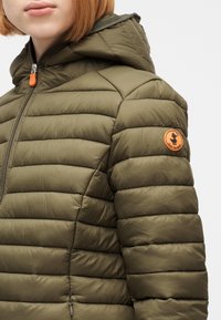 Γυναίκα που φοράει χακί μπουφάν καπιτονέ με κουκούλα και πορτοκαλί λογότυπο στο πάνω μέρος του μπράτσου, κλεισμένο με πορτοκαλί φερμουάρ.