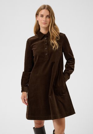 Femme portant une robe en velours côtelé marron foncé, longue jusqu'aux genoux, à manches longues, avec un col boutonné et des bottines noires.