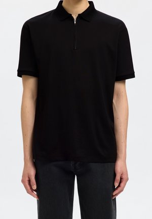 Homme portant un polo noir uni à manches courtes avec une fermeture éclair devant, associé à un pantalon noir, sur un fond clair uni.