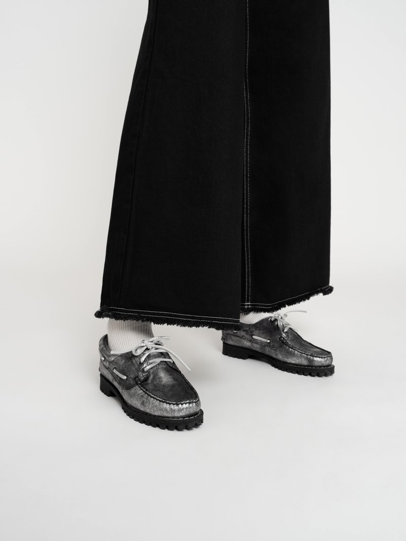 Schwarze Denim-Hose mit weitem Bein und ausgefransten Säumen, kombiniert mit grauen Lederschuhen mit strukturiertem Finish, runder Zehenform und weißen Schnürsenkeln.