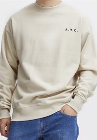 Osoba w beżowym swetrze z napisem "A.R.C." i niebieskich dżinsach, z jedną ręką w kieszeni, stojąca na tle białej ściany.