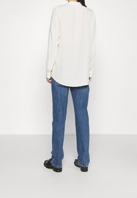 Maglietta crema a maniche lunghe con orlo arrotondato, abbinata a pantaloni di denim blu. La maglietta ha una texture liscia ed è progettata con maniche a bottoni.