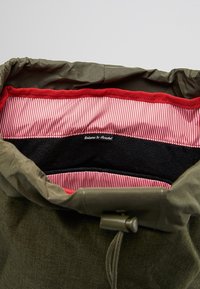 Zielona torba z materiału z zaciąganym otworem. Wewnątrz ma czerwone paski i miękką czarną podszewkę, z umieszczonym na środku logo marki u góry.
