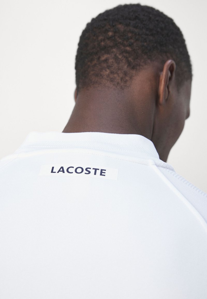 Polo blanc avec une finition texturée. Étiquette "LACOSTE" en bleu marine cousue à l'arrière. Col arrondi et manches courtes.