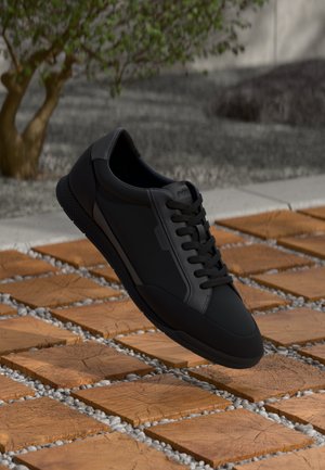 Fekete sneaker sima bőr felsőrésszel, kerek orral és fűzős dizájnnal, egy texturált fa és kavicsos felület előtt.