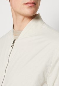 Lekka kremowa kurtka bombowiec z delikatnym wzorem w kratę, z zamkiem błyskawicznym i stójką. Miękka tkanina o gładkiej fakturze.