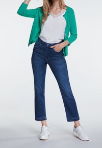 Grüne Strickjacke, weißes Oberteil mit Spitzenbesatz und dunkelblaue, kürzere Jeans. Die Turnschuhe sind weiß mit grauen Akzenten. Ein einfaches und klassisches Freizeit-Outfit.