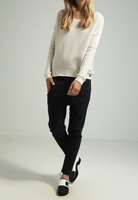 Strickpullover in Creme, lässiger Schnitt; schwarze, schmal zulaufende Hose; schwarz-weiße Schuhe mit Schnallendetail. Der Hintergrund ist ein einheitliches hellgrau.