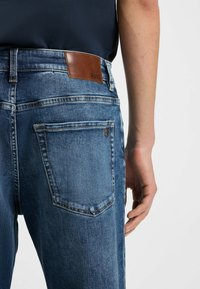 Jeans azul oscuro con una textura suave, que cuentan con un parche trasero de cuero marrón, costuras visibles y un diseño de bolsillo trasero sencillo.