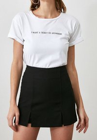 T-shirt blanc avec texte noir indiquant « JE VEUX UN BILLET POUR N’IMPORTE OÙ », associé à une mini-jupe noire avec des fentes sur les côtés et un tissu lisse.