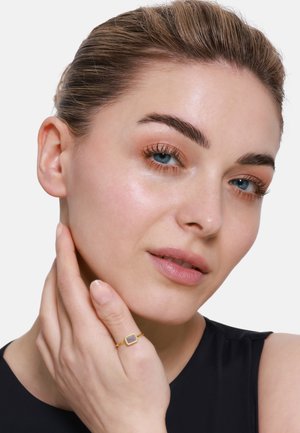 Goldring mit einem quadratischen Stein, der an einer Hand getragen wird, die das Gesicht berührt. Das Model hat glatte Haut, leichtes Make-up und ein schwarzes Oberteil. Heller Hintergrund.