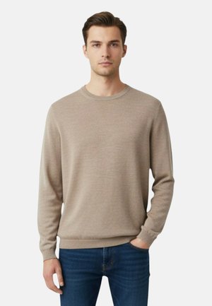 Uomo con maglione beige a girocollo e jeans blu, in piedi con una mano in tasca davanti a uno sfondo semplice.
