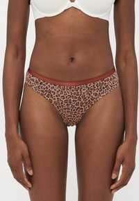 Woman wearing leopard print bikini-style briefs with rust-colored waistband and a white bra, standing with arms relaxed.