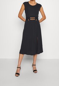 Černé midi šaty s krátkými rukávy, opaskem v pase s dvěma sponami a bočním rozparkem. Hladký materiál a volný střih. Nosené s černými sandály.