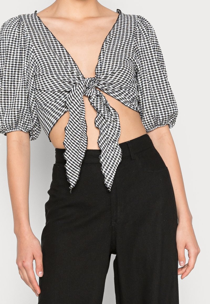 Femme portant un crop top vichy noir et blanc à manches bouffantes noué en nœud devant, associé à un pantalon noir taille haute.
