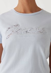 T-shirt di cotone azzurro chiaro con un logo "Guess" decorato con pizzo bianco e perle, riportante la scritta "Los Angeles" sotto, design a girocollo.